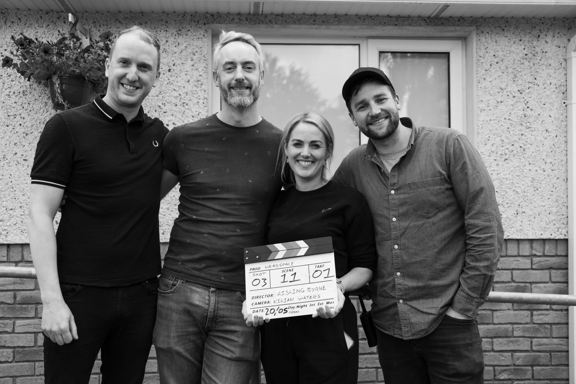 Black and white photo of four people standing outside in front of a brick wall, smiling, with one holding a film slate that has production information written on it.