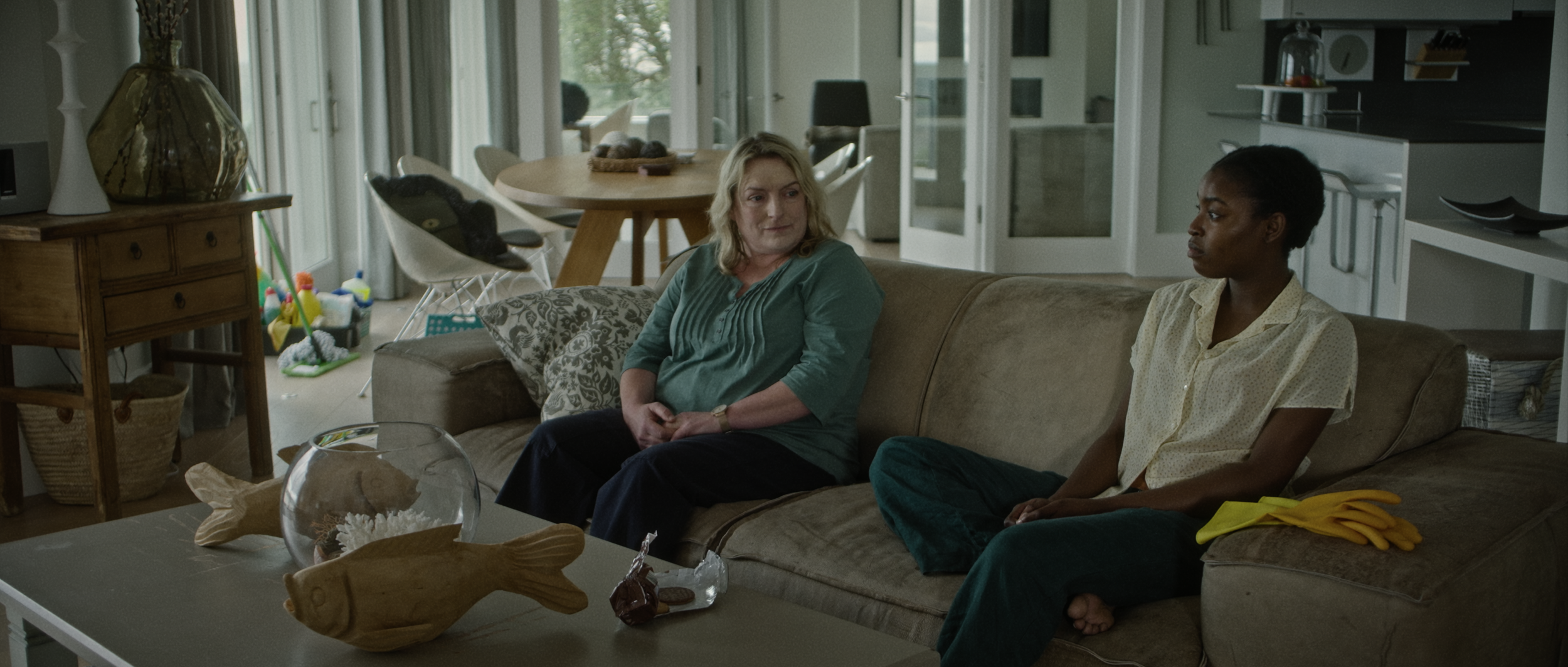 Two women sitting on a beige couch in a living room, engaged in conversation. One woman has blonde hair and is wearing a teal top, the other woman has dark hair and is wearing a yellow blouse.