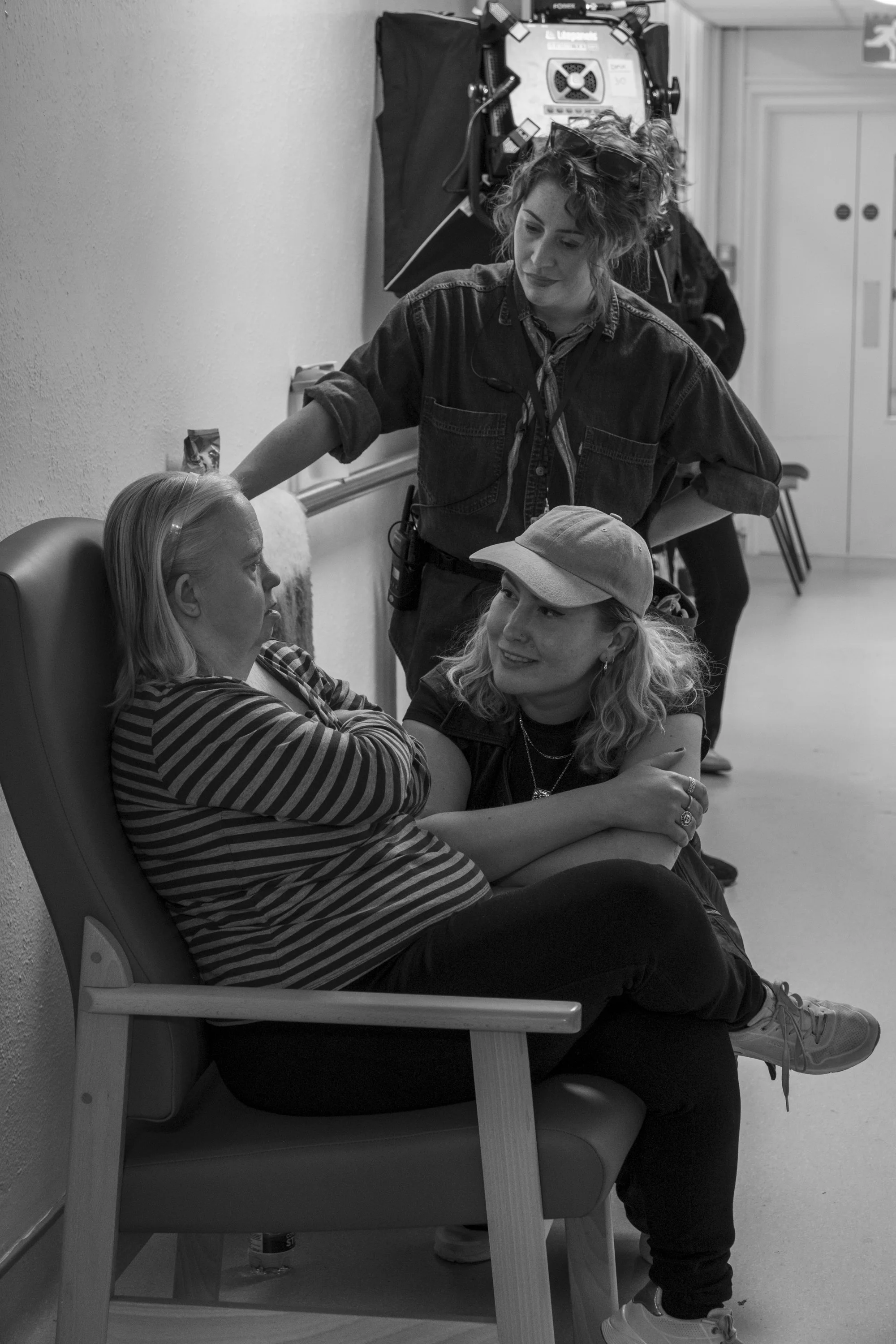 A woman with blonde hair and a striped shirt sitting on a chair with her arms crossed, talking with a woman with curly hair, a baseball cap, and a tank top. The curly-haired woman is sitting on a chair with legs crossed, smiling, and holding the blon