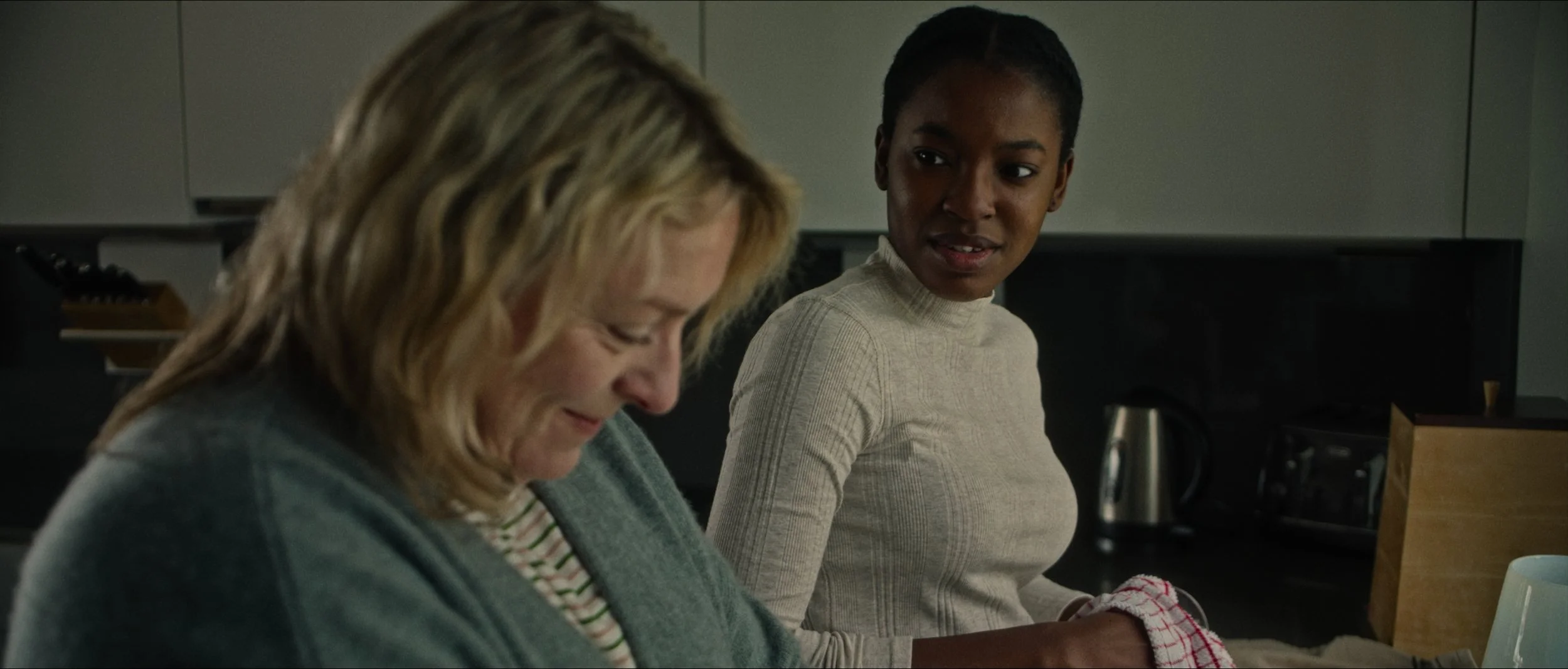 Two women in kitchen, one with blonde hair looking down, other with dark hair looking at her, near kitchen appliances and counter.