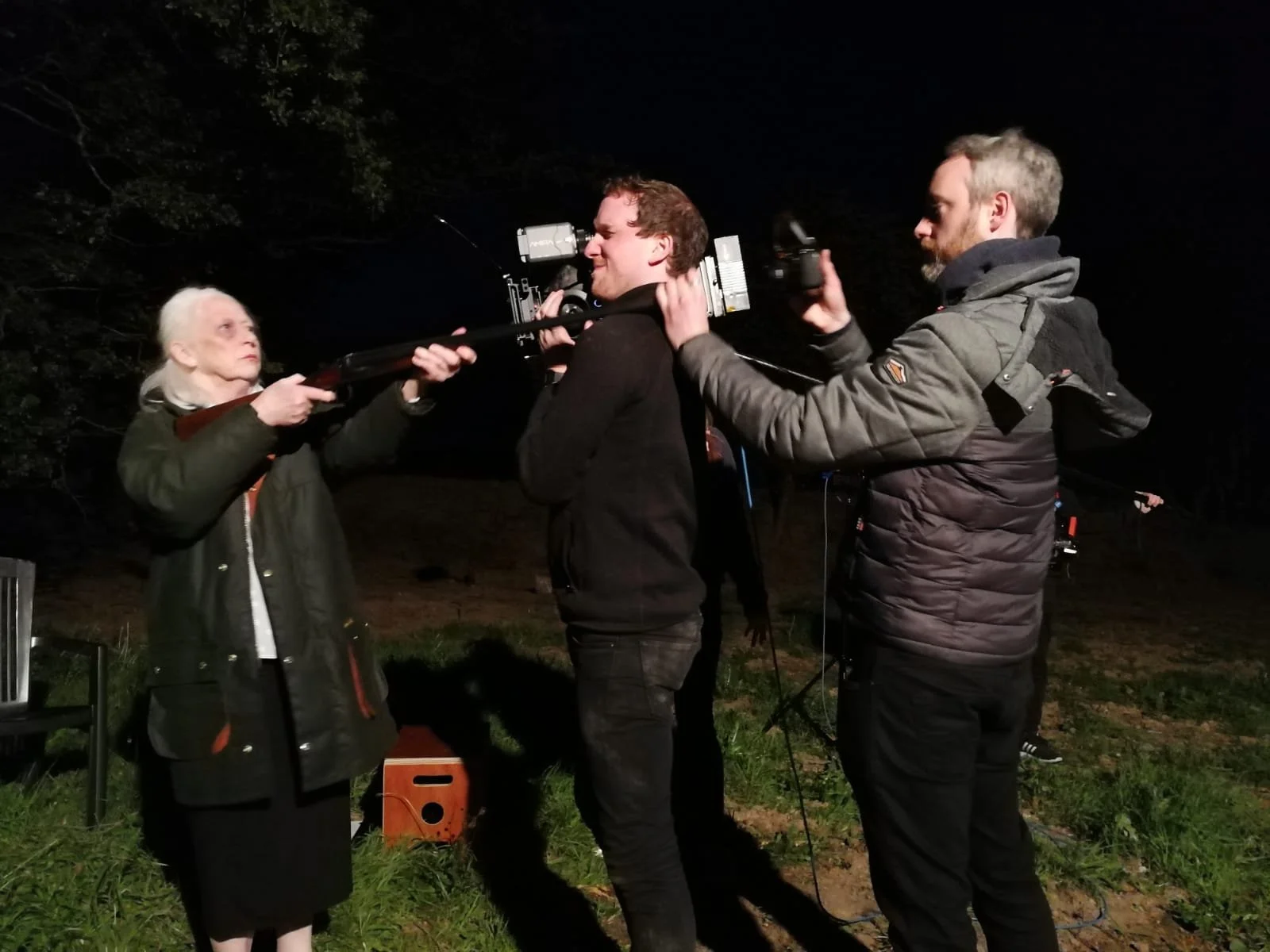 A woman is holding a rifle, while another man adjusts a camera on a tripod to film the scene at night outdoors.