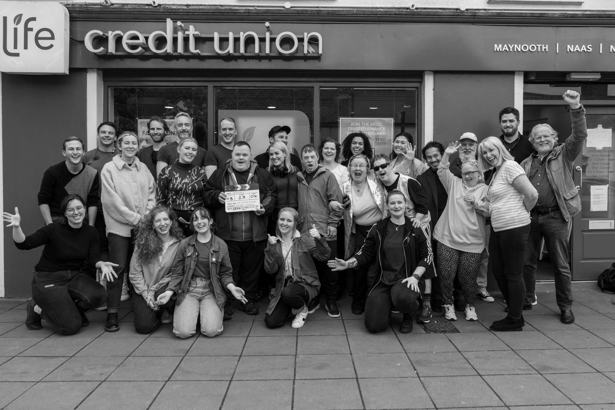 Group of diverse people smiling and posing in front of a credit union building, with some making gestures and one holding a clapperboard.