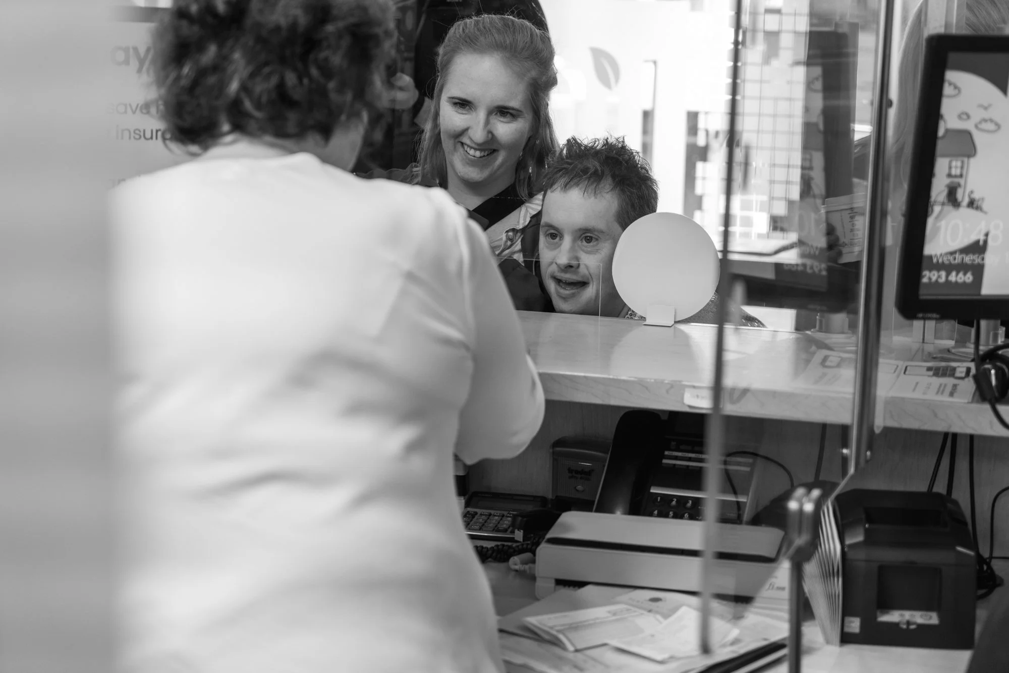 A woman smiling at a man during a visit to a healthcare or office reception area, with a desk, computer, and informational signage visible.