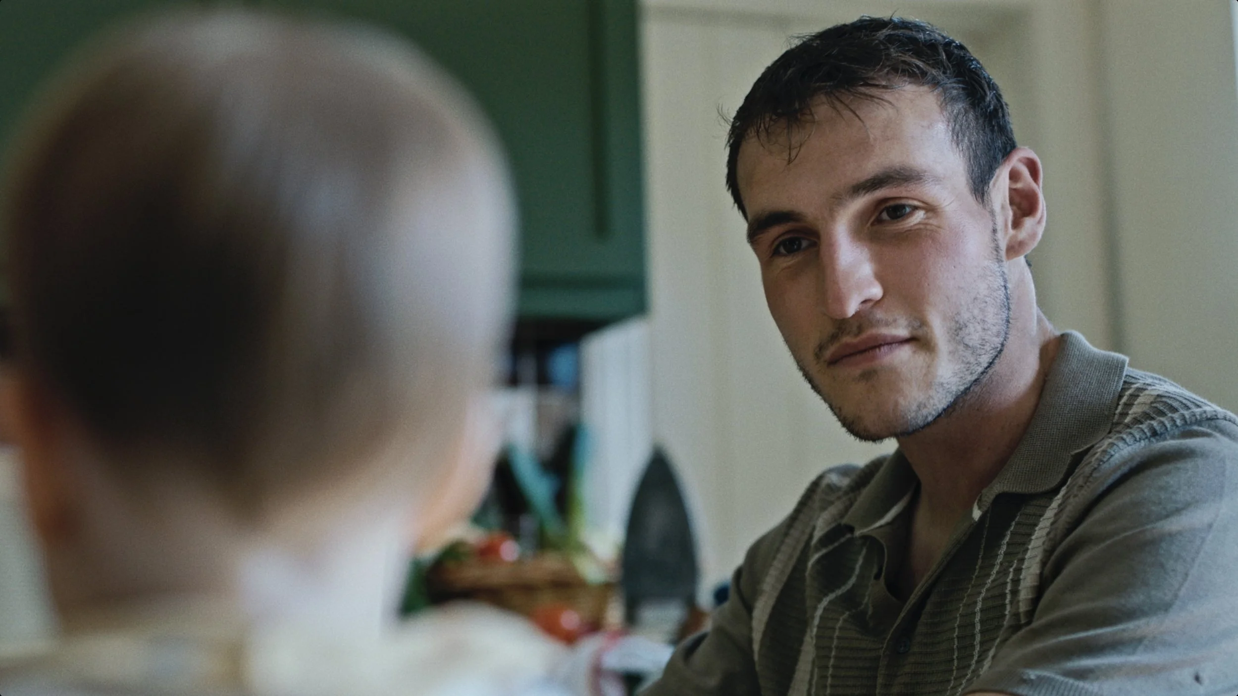 A young man with dark hair and a beard looks at a blurred person in the foreground, with a home setting in the background.