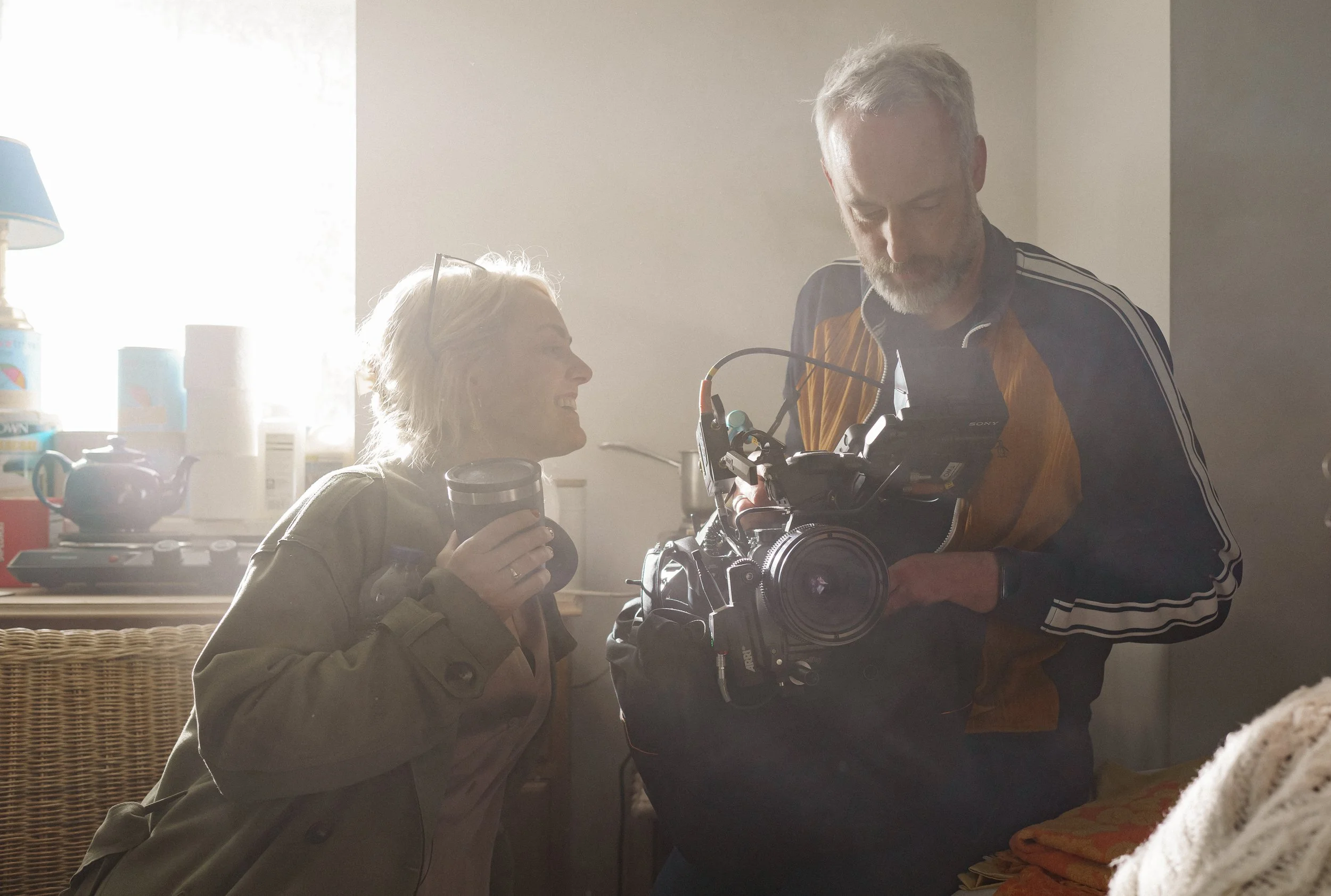 A man and a woman on a film set, with the man operating a camera and the woman holding a cup, sharing a moment in a cozy kitchen.