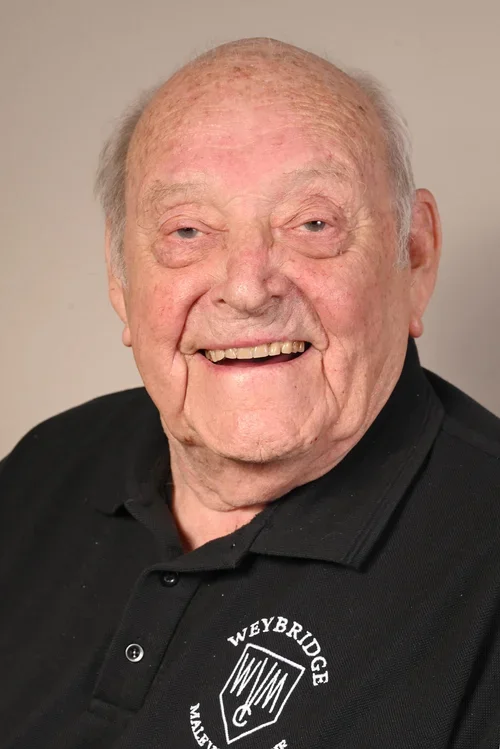 An elderly man with a big smile, showing his teeth, wearing a black polo shirt with a "Weybridge" logo.