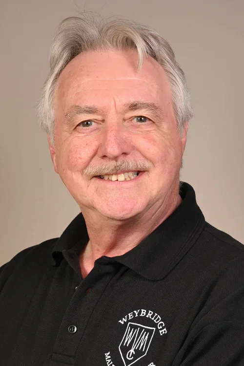 A smiling older man with gray hair, mustache, and blue eyes, wearing a black polo shirt with a logo that reads "Weybridge".