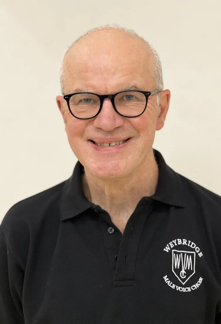 A middle-aged man smiling, wearing glasses and a black polo shirt with a logo that reads "Weybridge Male Voice Choir."