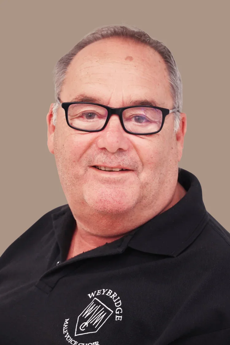 A middle-aged man with glasses, wearing a black polo shirt with the logo 'Neybridge Male Voice Choir,' smiling against a plain background.