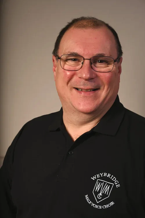 A man with glasses and short hair, smiling, wearing a black polo shirt with a Weybridge Male Voice Choir logo.