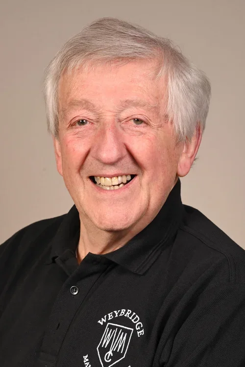 An elderly man with gray hair smiling, wearing a black polo shirt with a Weybridge logo.