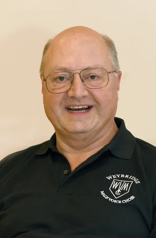 A middle-aged man with glasses and a bald head, smiling, wearing a black polo shirt with the logo of WBYBRIDGE Male Voice Choir.