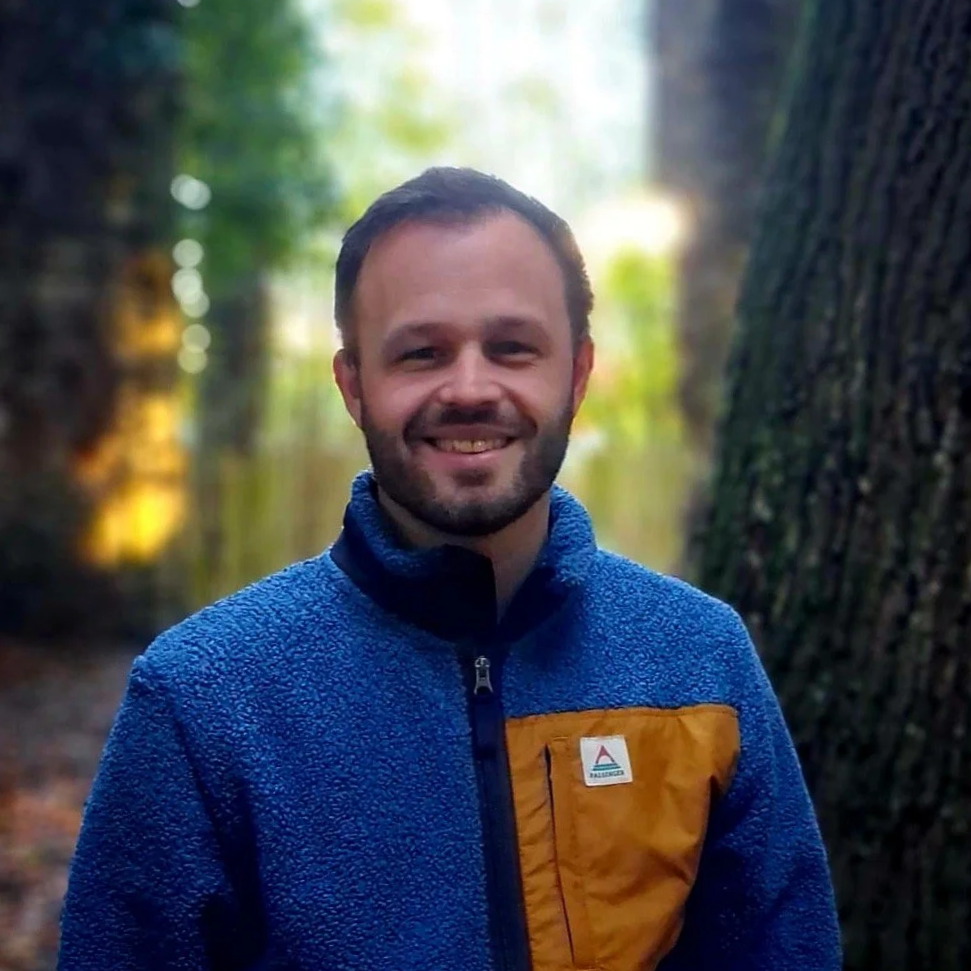 A man with short brown hair and a beard, smiling, standing in a forest with trees and sunlight in the background. He is wearing a blue fleece jacket with a brown chest pocket.