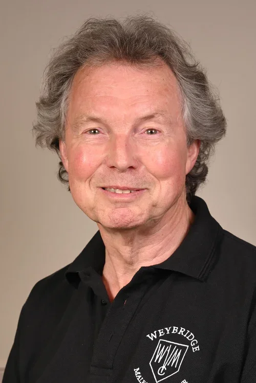 A middle-aged man with gray, curly hair wearing a black polo shirt with 'Weybridge' and a logo on it, smiling at the camera.