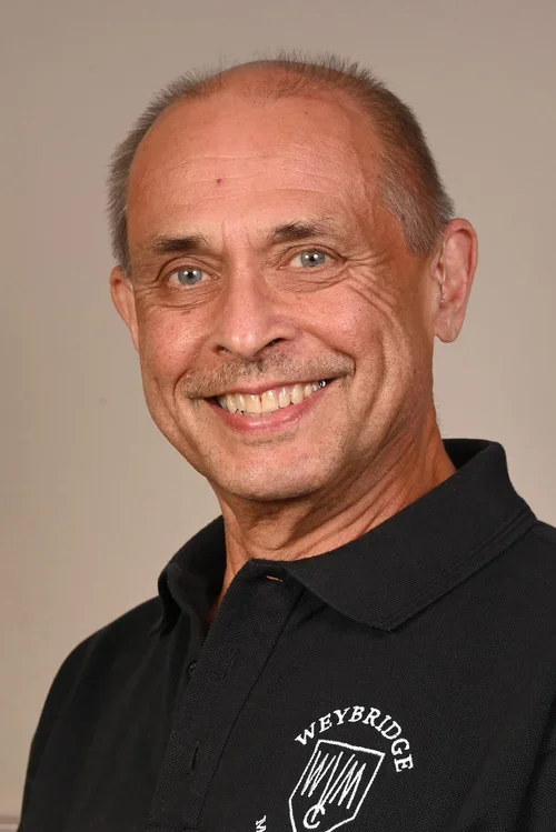 A smiling middle-aged man with short, thinning hair, wearing a black polo shirt with a logo and text that reads 'Weybridge' on the chest.