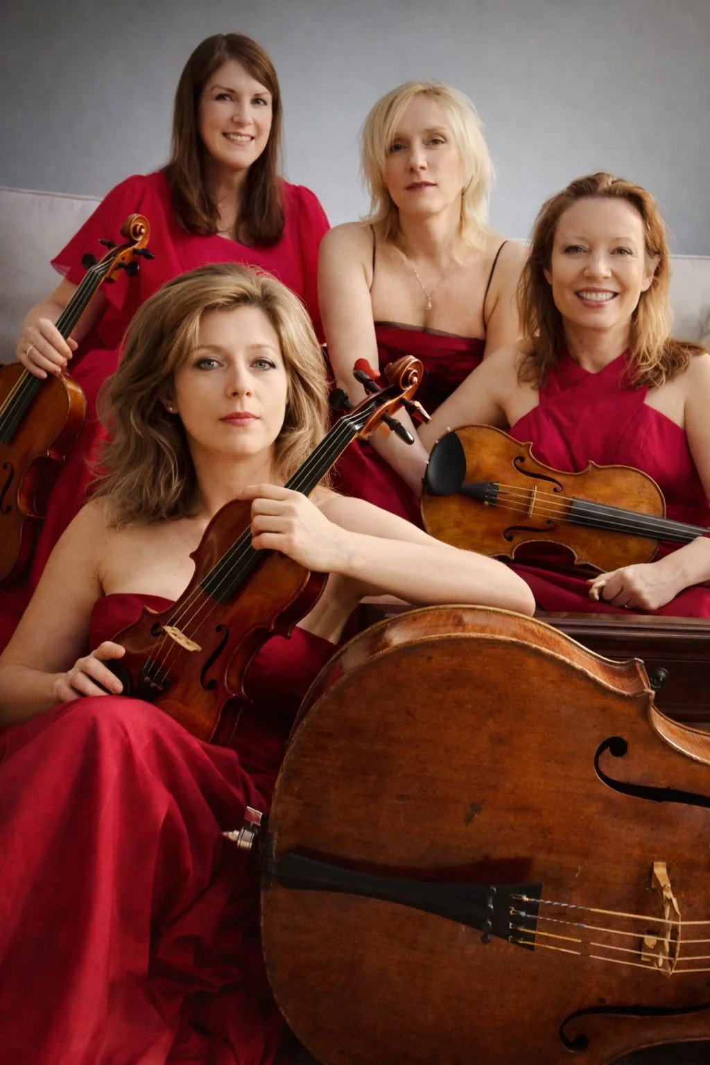 Group of four women holding violins, sitting on a couch, dressed in red dresses, with one woman in the front holding a violin, and a double bass in front of her.