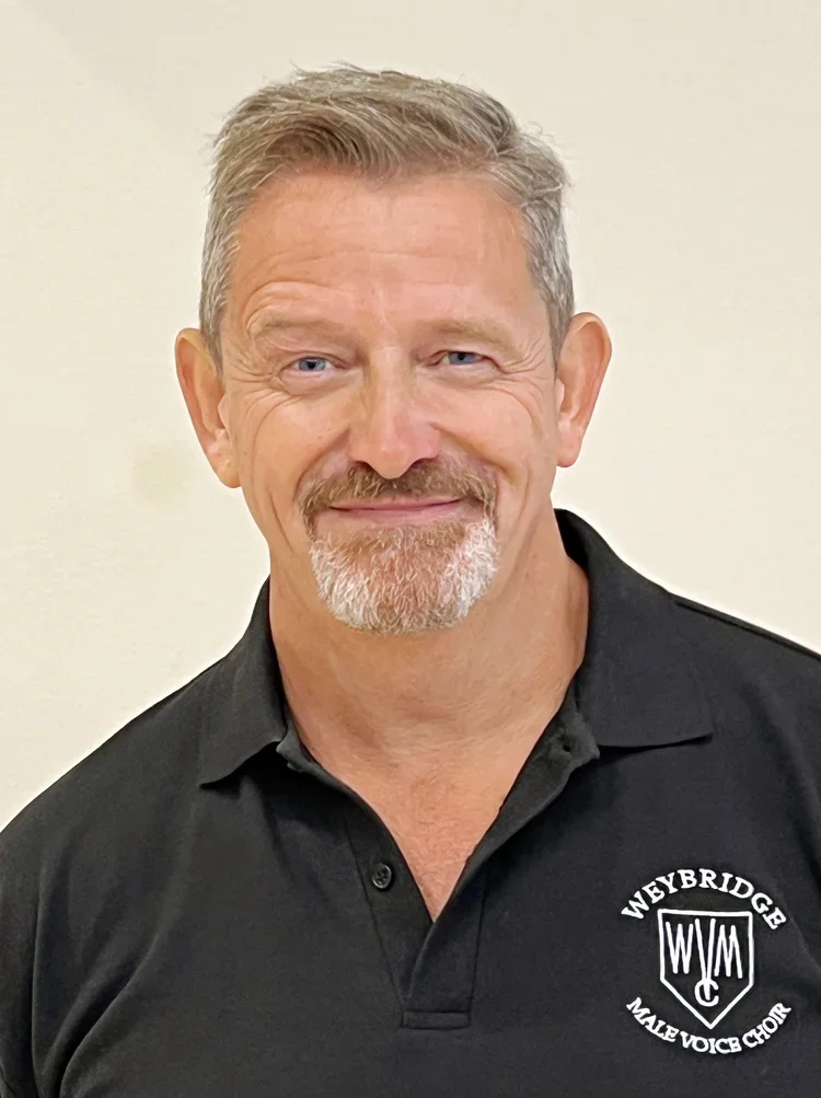 A man with gray hair and a beard wearing a black polo shirt with a logo for Weybridge Male Voice Chorus.