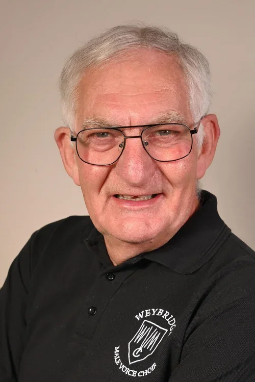 Senior man with gray hair and glasses wearing a black polo shirt with a Weybridge May Voice Choir logo, smiling at the camera.