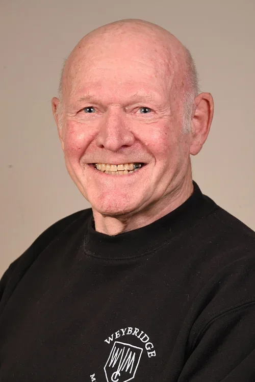 A smiling elderly man with blue eyes and a bald head, wearing a black sweatshirt with the word 'Weybridge' and a crest logo.