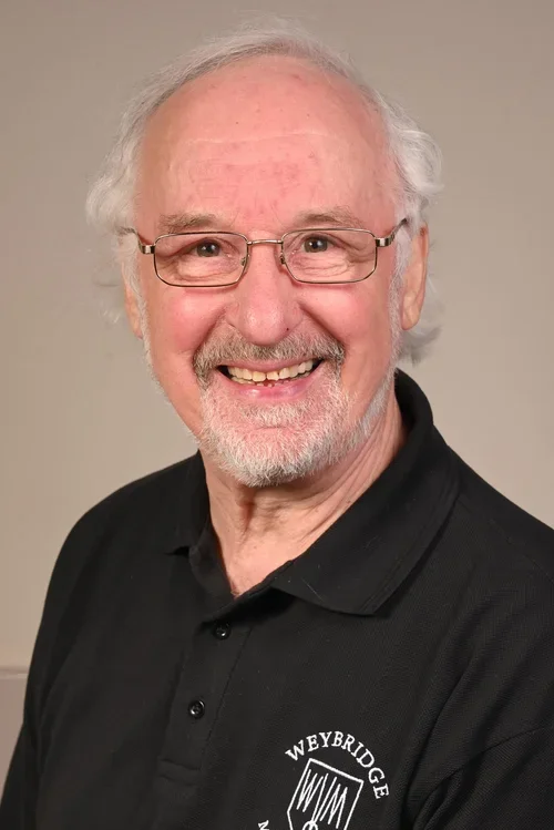 A smiling older man with white hair, glasses, and a beard, wearing a black polo shirt with a logo that says 'Weybridge' and 'WWM' on it.