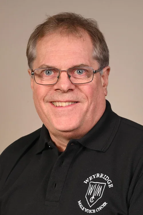A middle-aged man with glasses, smiling, wearing a black polo shirt with a 'Weybridge Male Voice Choir' logo.