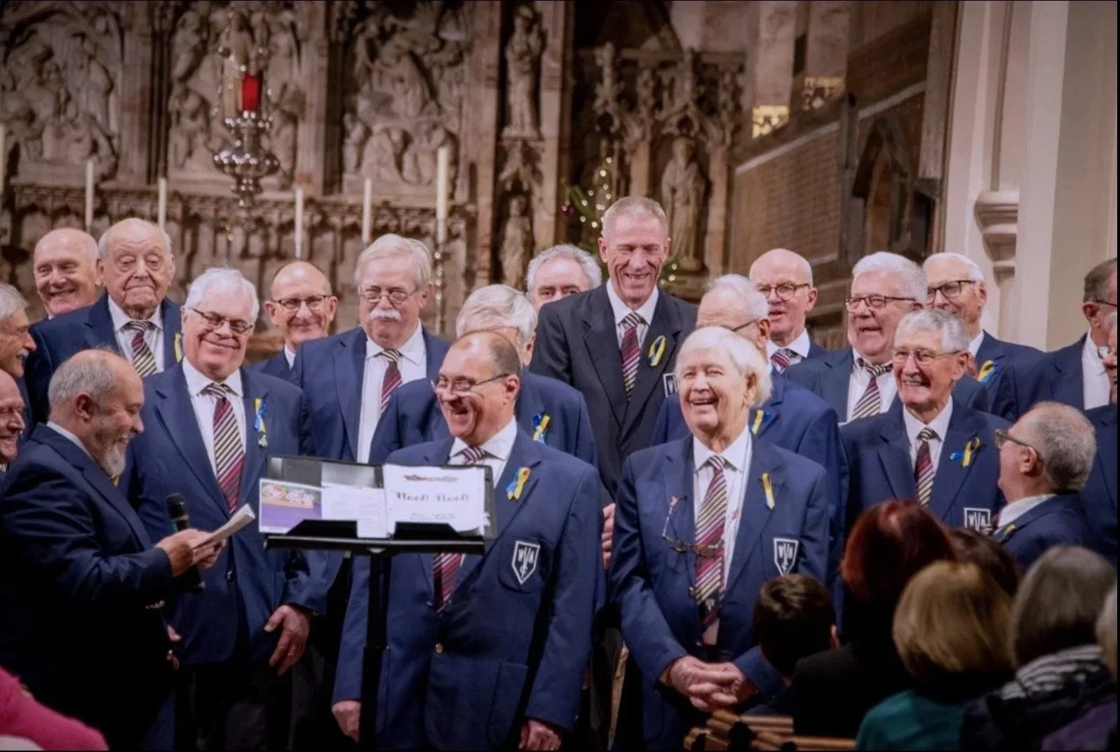 A group of men, mostly elderly, dressed in matching blue suits with yellow ribbons, singing or performing in a church with wooden carvings and candles in the background.
