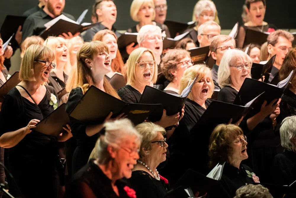 Group of mature men and women singing in a choir, holding black songbooks, wearing black clothing, some with glasses, performing passionately.