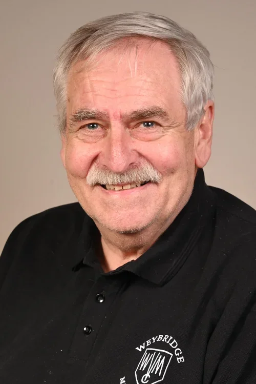 A smiling elderly man with gray hair and a mustache, wearing a black polo shirt with the Weybridge logo, against a neutral background.