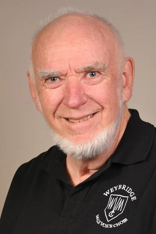 A smiling elderly man with a white beard and bald head, wearing a black polo shirt with the logo and text for Weybridge Masonic Choir.
