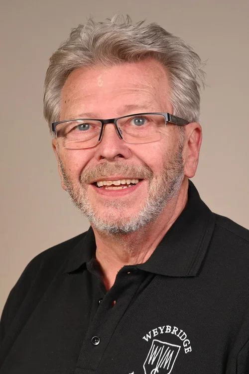 A smiling middle-aged man with glasses and gray hair, wearing a black polo shirt with a logo on it.
