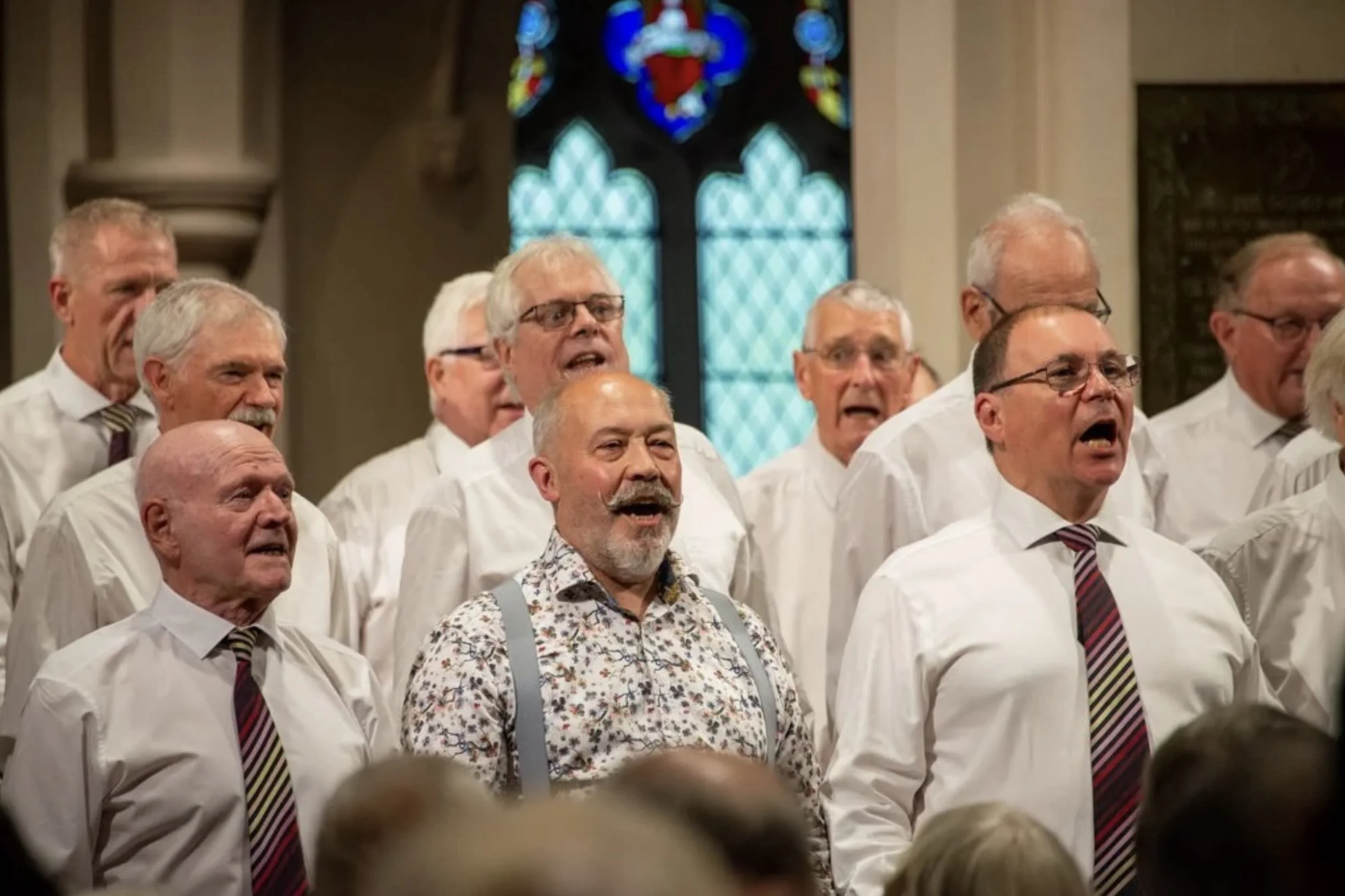 A men's choir singing in a church with stained glass windows in the background.