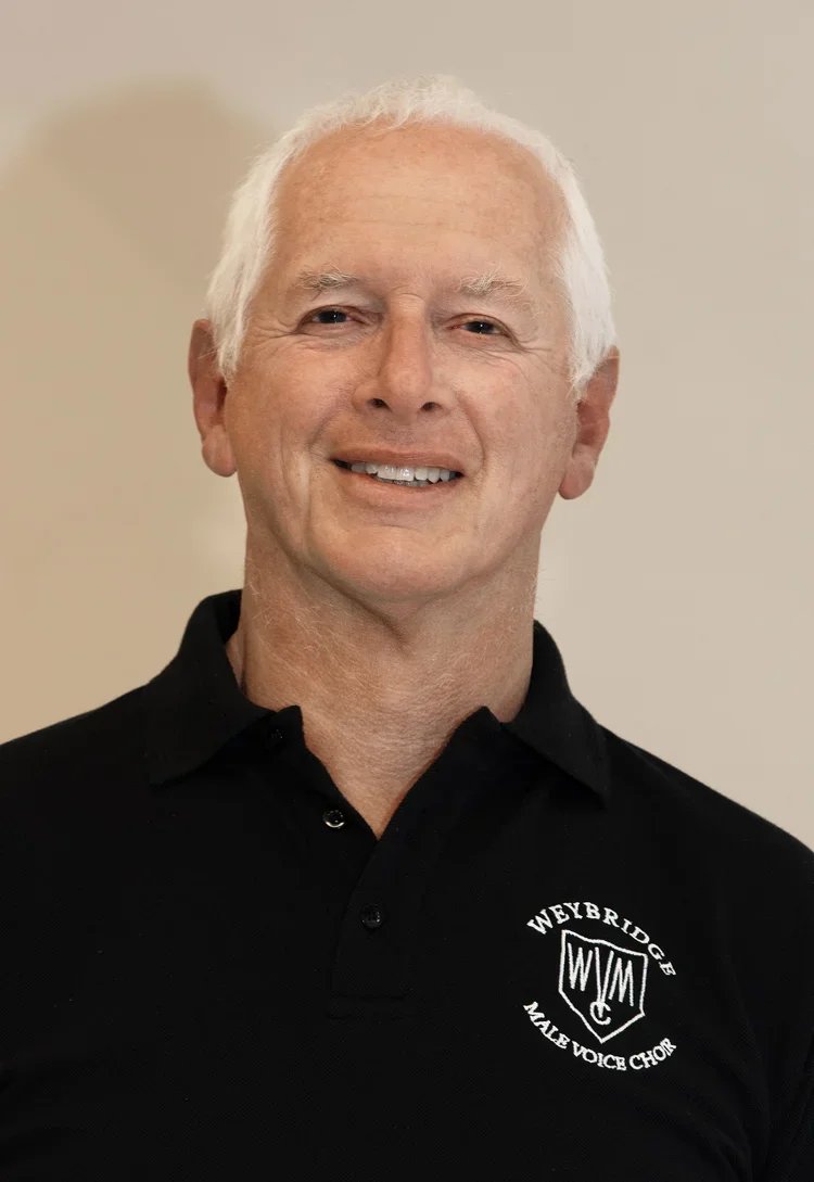 A smiling older man with white hair wearing a black polo shirt with a 'Weybridge Male Voice Chorus' logo.