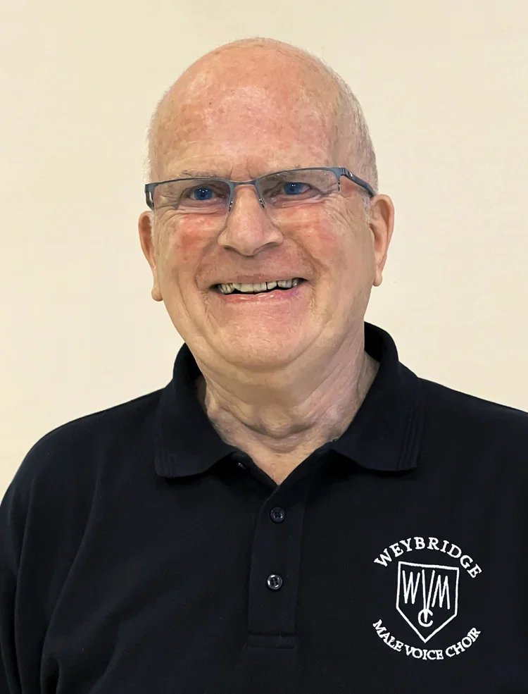 A smiling elderly man with glasses wearing a black polo shirt with a Weybridge Male Voice Choir logo.