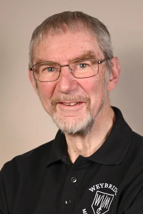 A middle-aged man with glasses, gray hair, and a beard smiling at the camera, wearing a black polo shirt with a Weybridge logo.