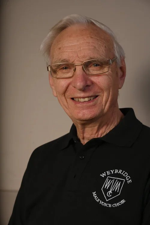 An elderly man with gray hair and glasses, smiling in a black collared shirt with a logo for Weybridge Male Voice Choir.