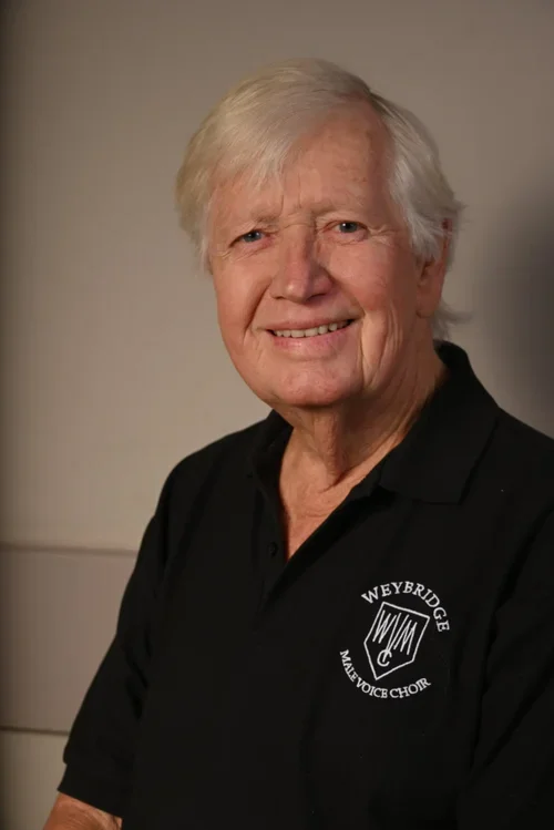 Older man with white hair smiling, wearing a black polo shirt with the logo of Weybridge Male Voice Choir.