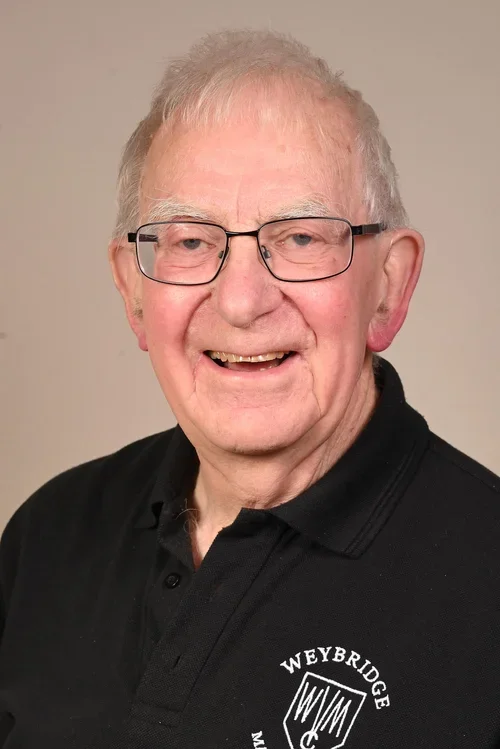 An elderly man with gray hair, glasses, and a wide smile, wearing a black polo shirt with the Weybridge logo.