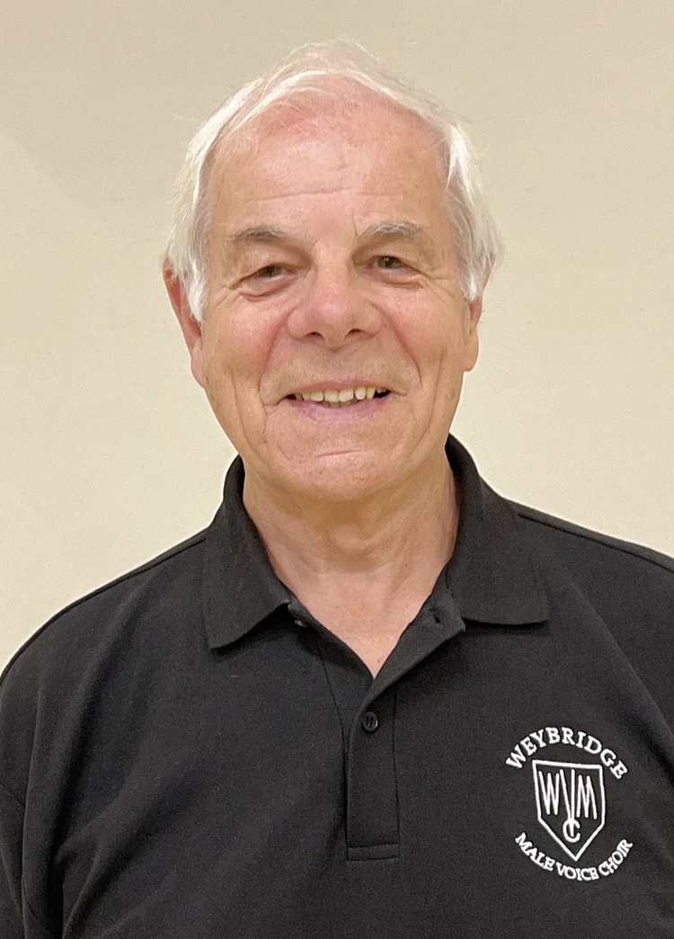 A senior man smiling, wearing a black polo shirt with the logo 'Weybridge Male Voice Choir' on the chest, standing against a plain beige background.