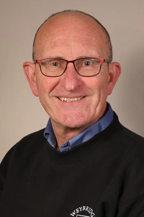 A middle-aged man with glasses, short light hair, and a friendly smile, wearing a blue collared shirt and a black sweater with a logo on it.