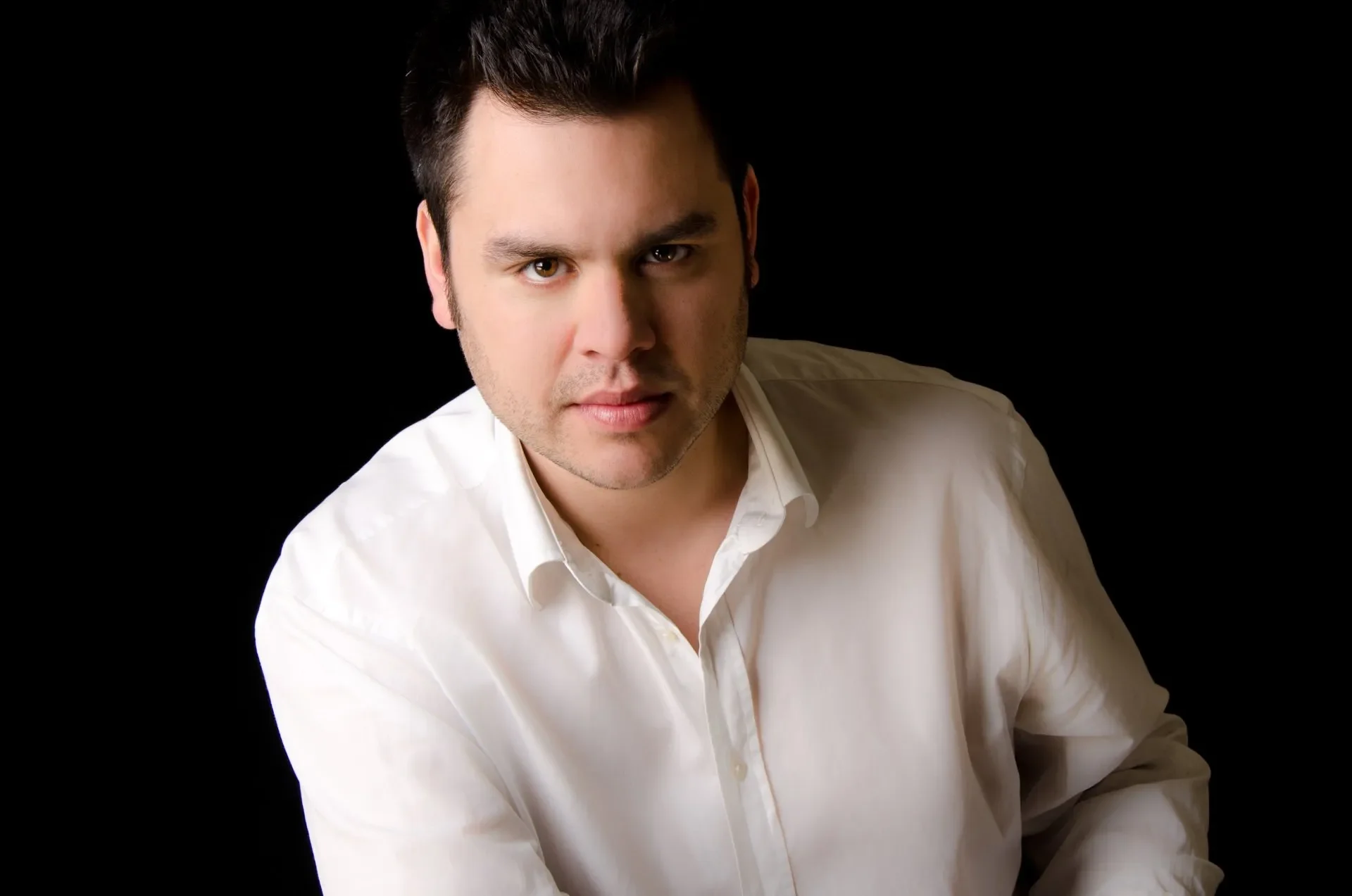 Portrait of a young man with dark hair, wearing a white dress shirt, looking confidently at the camera against a black background.