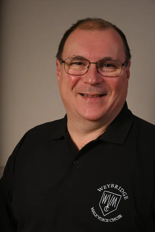 A middle-aged man with glasses, short brown hair, and a friendly smile, wearing a black polo shirt with a Weybridge Male Voice Choir logo.