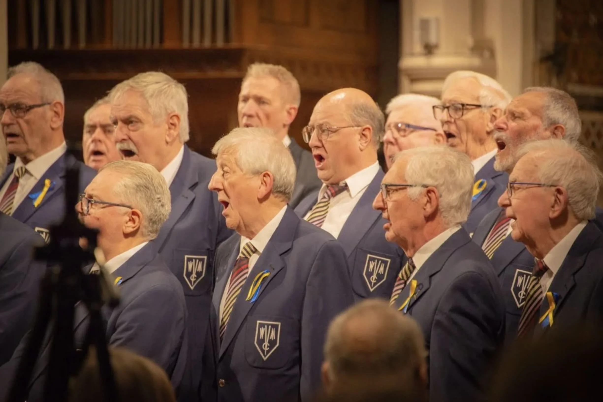 A group of elderly men wearing matching navy blue suits, white shirts, and striped ties, singing or reciting in a formal setting with wooden shelves or bookcases behind them.