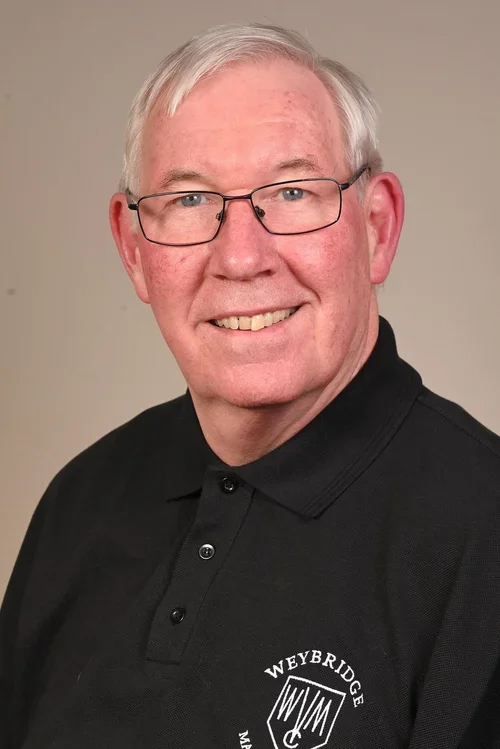 A smiling older man with gray hair and glasses wearing a black polo shirt with the Weybridge logo.