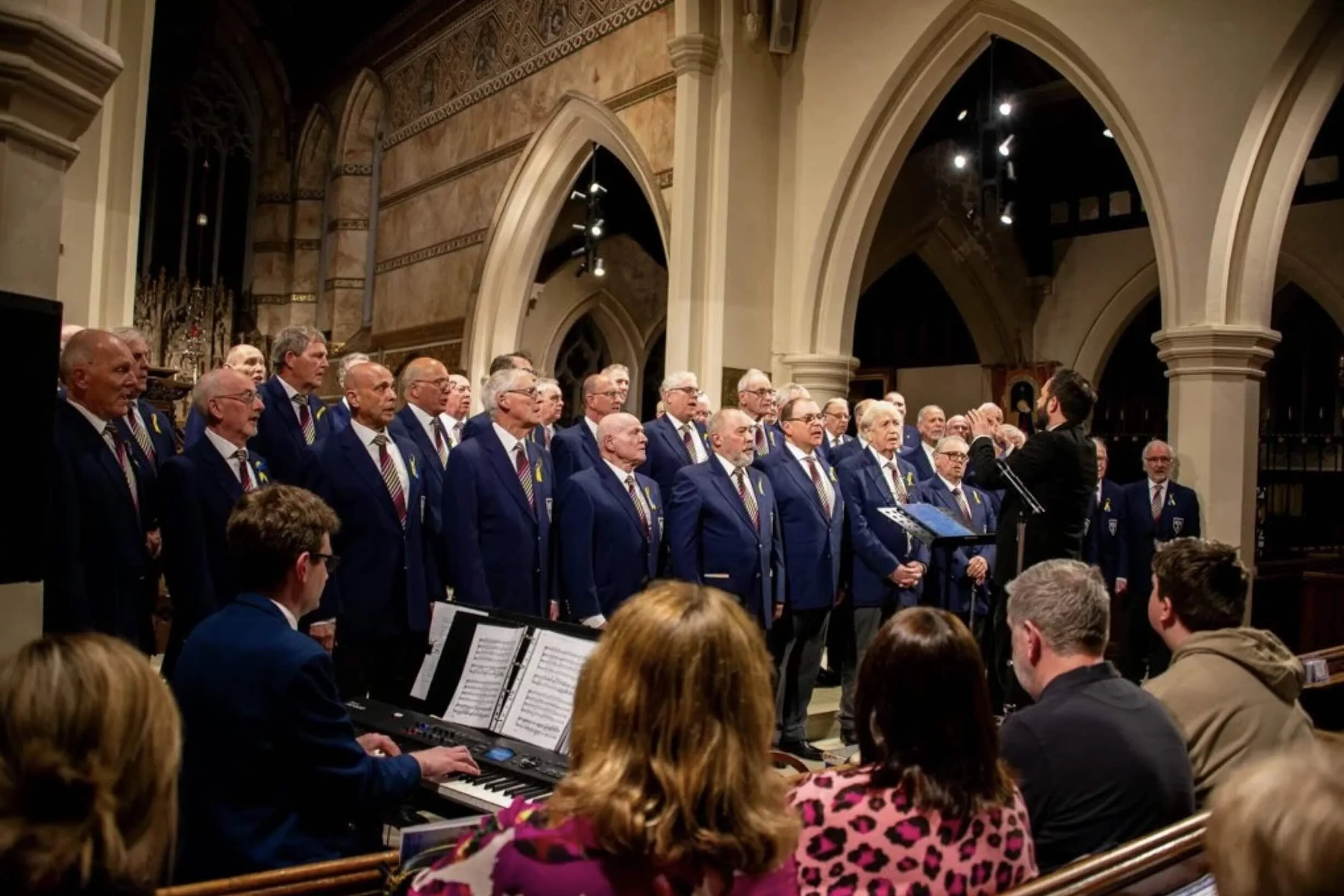 A men's choir dressed in blue blazers and striped ties performing in a church, led by a conductor, with an audience seated in front.
