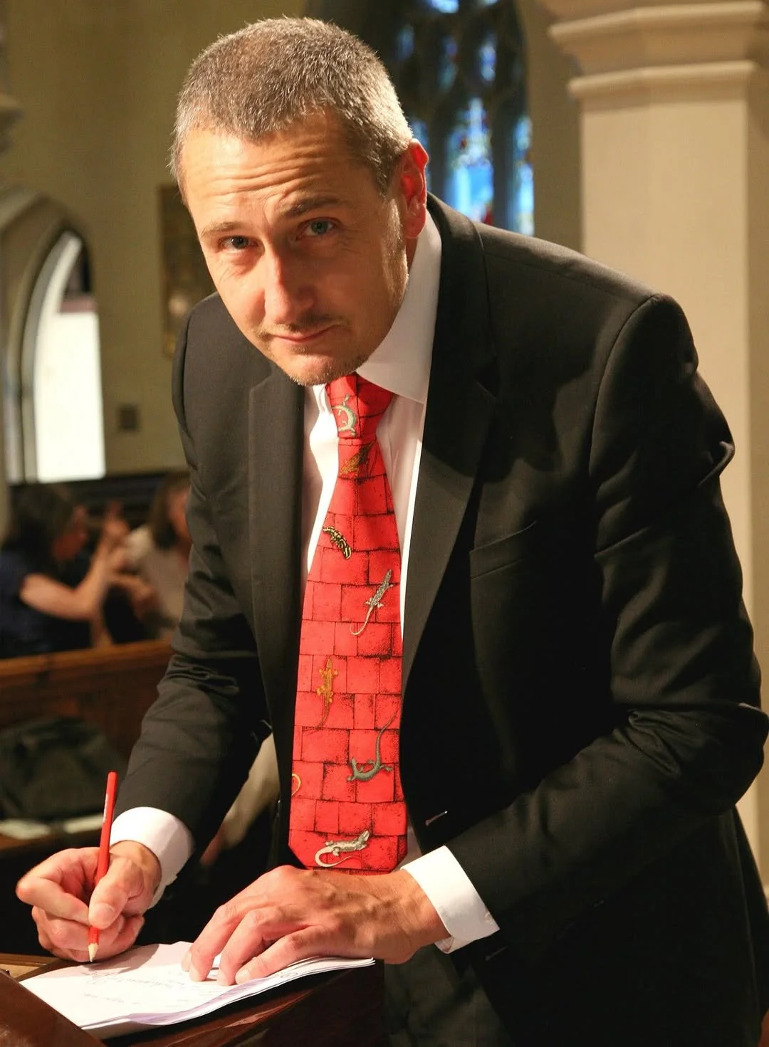 A man in a black suit and red tie with colorful lizard and snake designs, signing a book.
