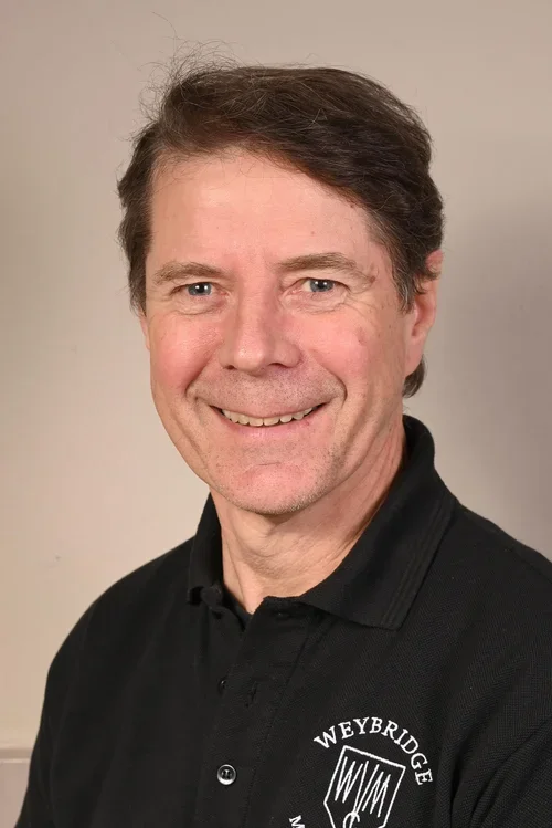 A man with dark hair and light skin wearing a black polo shirt with an embroidered logo that says Weybridge, smiling at the camera against a plain background.
