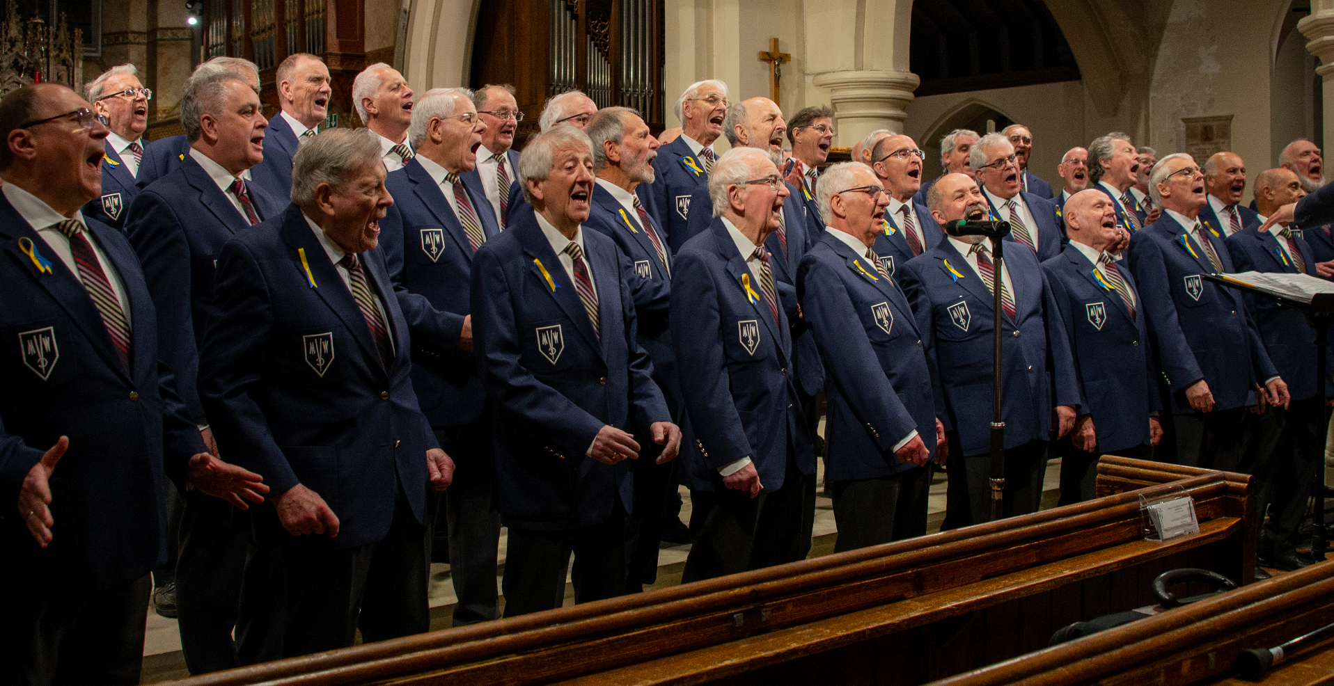 Group of elderly men singing together in a church, wearing matching blue blazers with patches and yellow ribbons.
