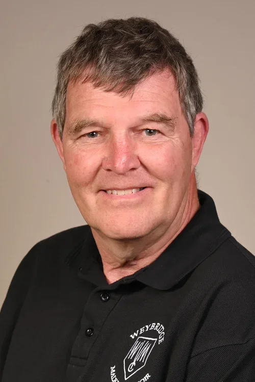 A middle-aged man with short, gray hair and blue eyes smiling at the camera, wearing a black polo shirt with a logo on the chest that reads "Weybred" and has a shield icon.