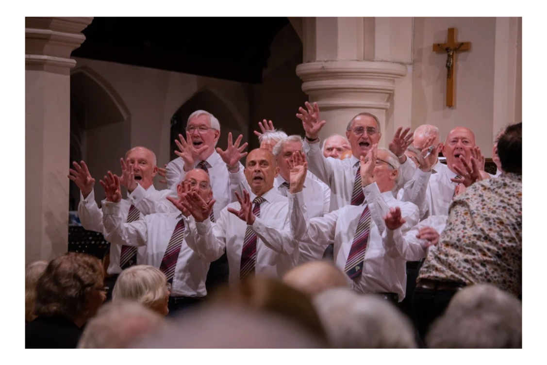 A group of men singing or performing in a church, with some raising their hands, dressed in white shirts and striped ties, with a crucifix on the wall behind them.