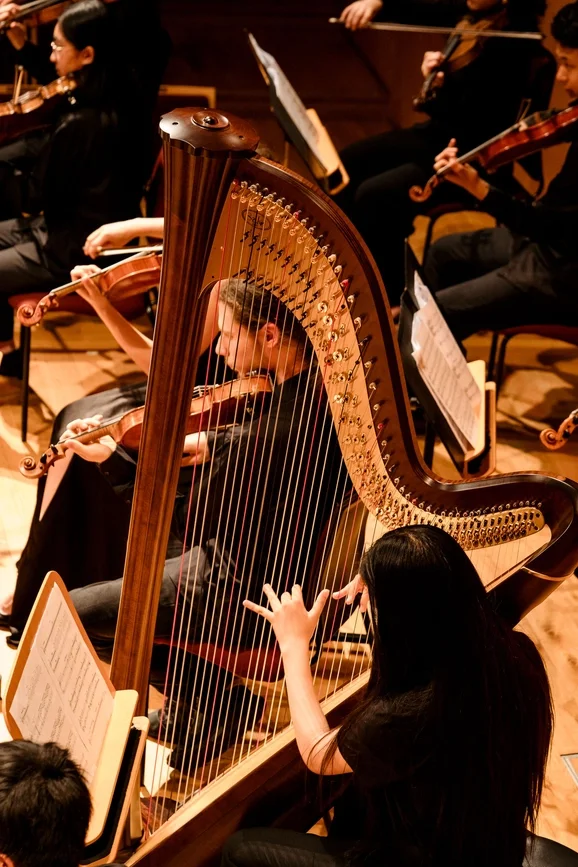 A young woman is conducting or guiding a harpist during a classical orchestra performance, with other musicians playing violins and cellos in the background.
